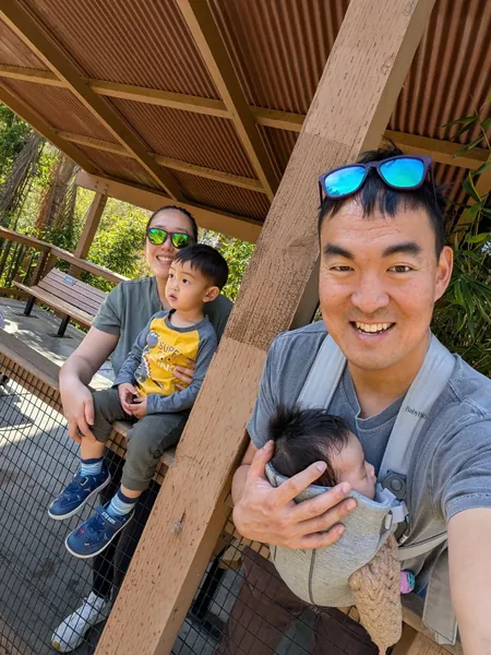 Jonathan on a shaded walkway with Maggie in a baby carrier, Tina and Everest on a bench behind them