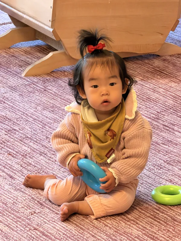 Maggie sitting on the rug in a knitted sweater, holding a blue stacking ring